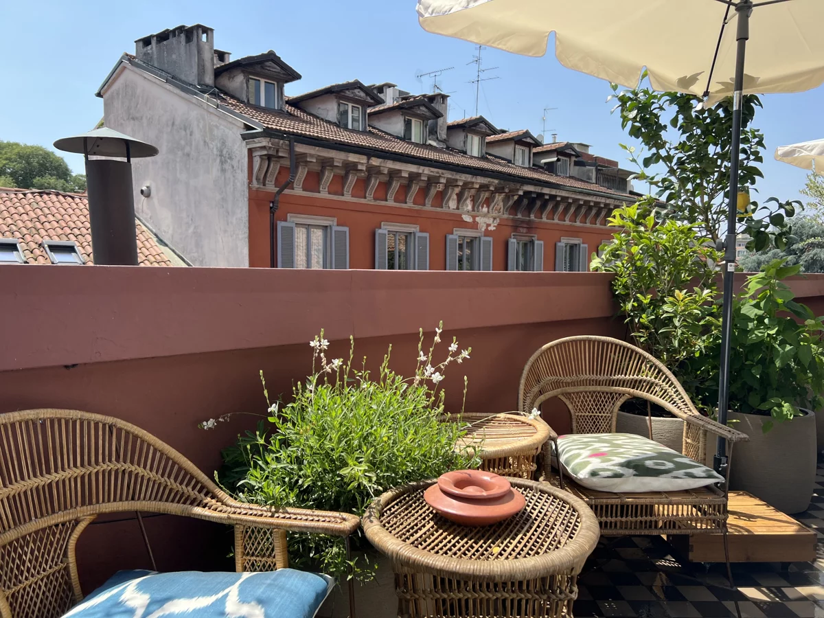 Terrazzo con poltrone in rattan, tavolino basso e piante in vaso, affacciato sui tetti e sulle facciate di Milano
