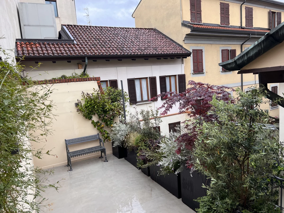 Terrazzo con panchina e fioriere scure piene di arbusti, tra le case e i tetti di Milano